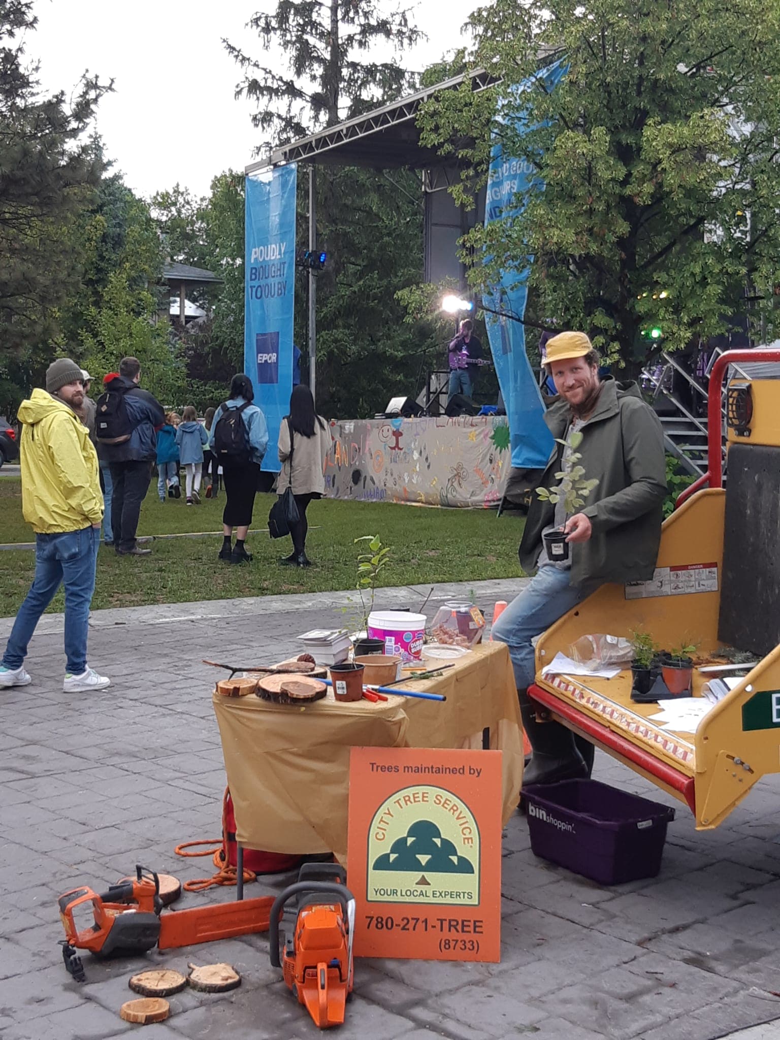 City Tree Service team member at a community education booth at Highlandia, with tree saplings, wood cookies, and a Trees Maintained By City Tree Service sign